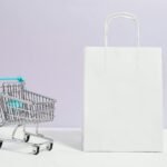 Minimalist image of a white shopping bag and mini cart on a soft pastel background.